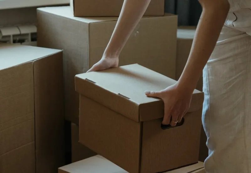 Une personne soulève un carton à couvercle devant une pile de cartons